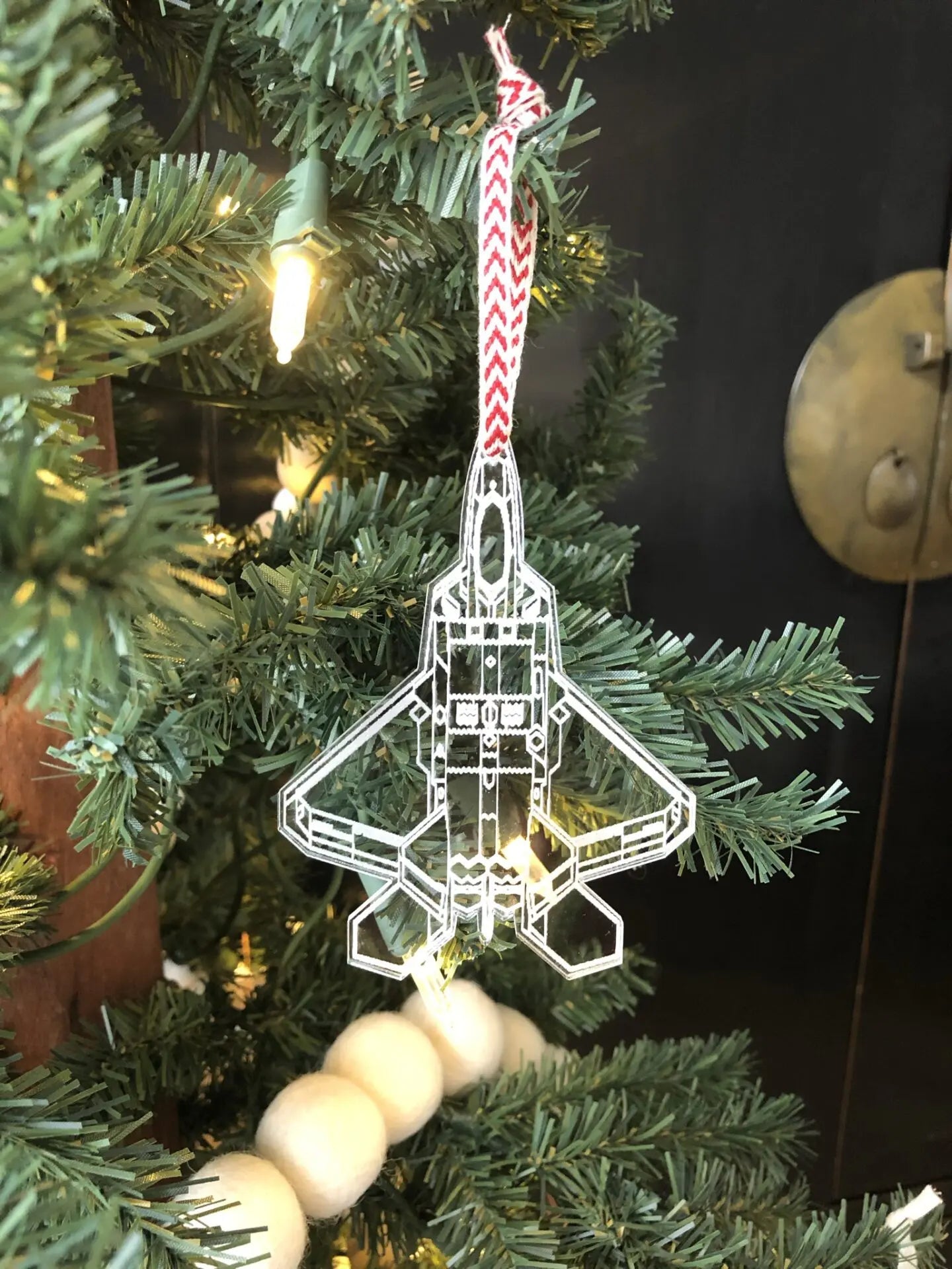 A clear acrylic Christmas ornament shaped like a fighter jet hangs from a red and white chevron striped ribbon on a decorated Christmas tree with warm lights.