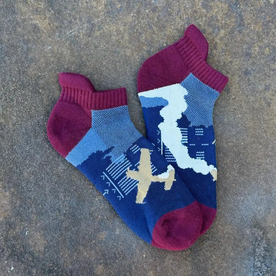A pair of low-cut ankle socks with a blue, navy, and burgundy knit pattern featuring a cityscape, an airplane, and a map design, laid flat on a textured gray surface.